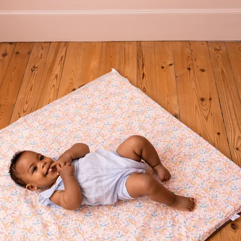 Bébé qui suce son doigt allongé sur le tapis rose pâle à petites fleurs posé en losange sur un parquet, vue plongeante