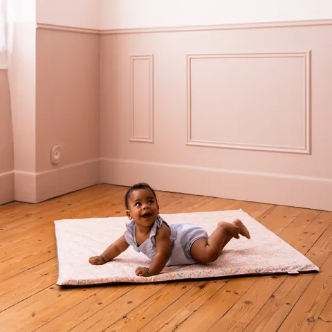 Bébé sur le ventre qui sourit sur le tapis Poésie, parquet et murs roses, plan large