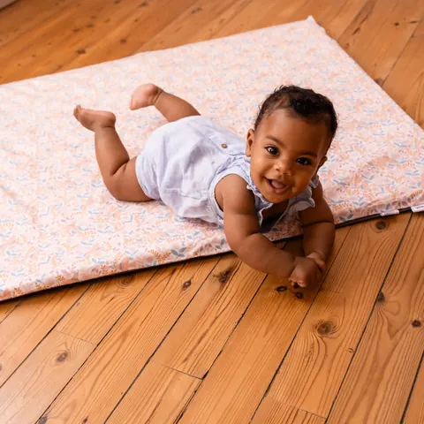 Bébé souriant sur le ventre au bord du tapis Poésie, parquet en bois