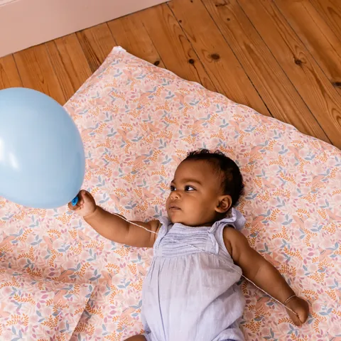 Bébé allongé qui tient un ballon bleu sur le tapis Poésie posé sur un parquet