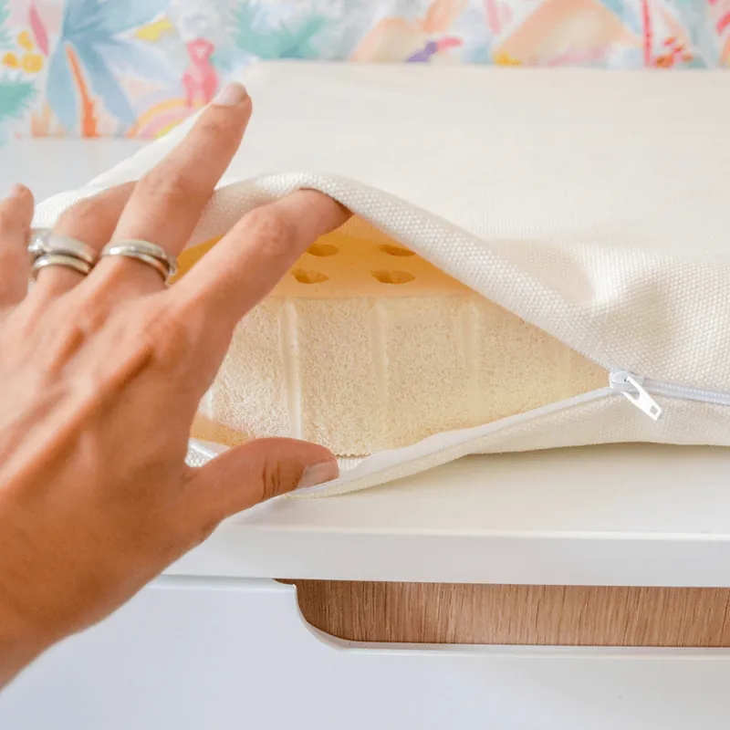 Une main ouvre la fermeture éclair de la housse en coton écru d'un matelas à langer et laisse voir la mousse de latex perforée