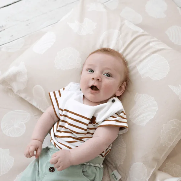 Un bébé en tee-shirt rayé marron, allongé sur un grand oreiller imprimé de coquillages, regarde l'objectif la bouche ouverte