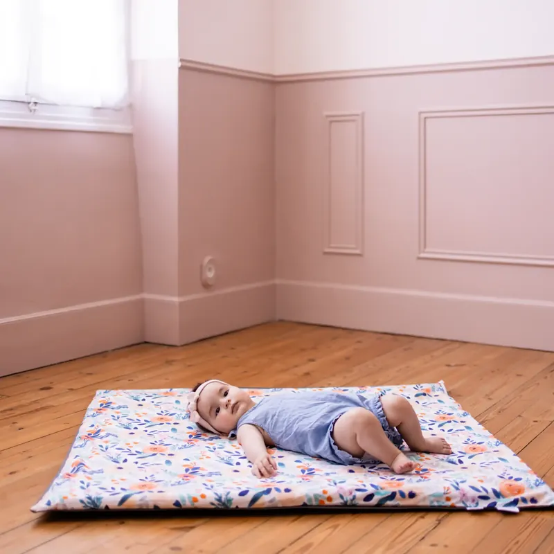 Un bébé en barboteuse bleue allongé sur le dos sur un tapis d'éveil fleuri, posé sur le parquet d'une chambre aux murs roses