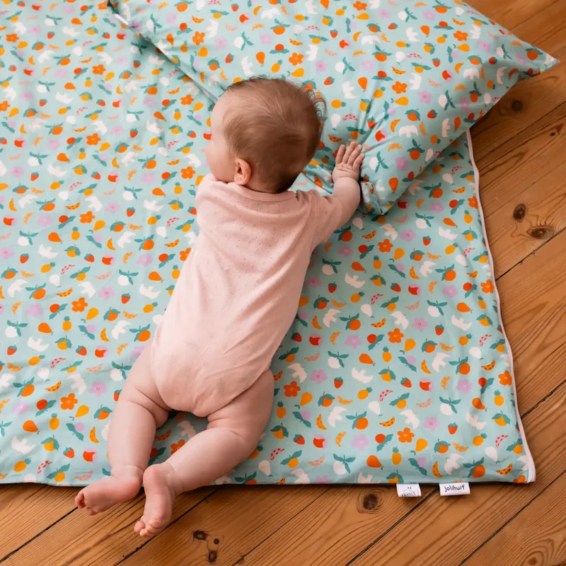 Un bébé en body rose, à plat ventre sur un tapis d'éveil imprimé de fruits et de fleurs, s'appuie sur le coussin assorti, sur un parquet