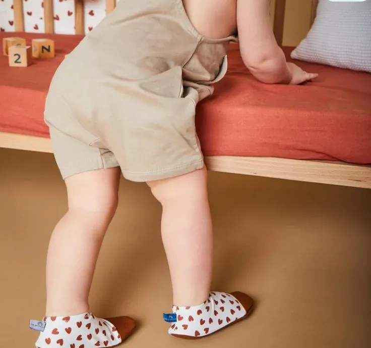 Un enfant debout contre un lit, chaussé de chaussons souples blancs à petits cœurs rouges