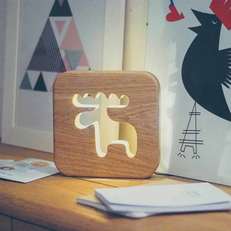 Une veilleuse en bois allumée, découpée en forme de caribou, posée sur un meuble devant des affiches illustrées