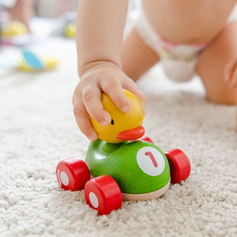 Une main de bébé pousse une petite voiture en bois verte à roues rouges, conduite par un canard jaune, sur un tapis moelleux