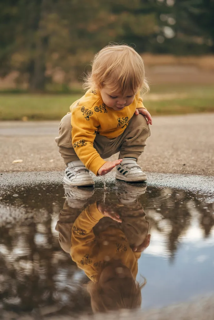 Un enfant accroupi au bord d'une flaque d'eau en touche la surface, son reflet dans l'eau