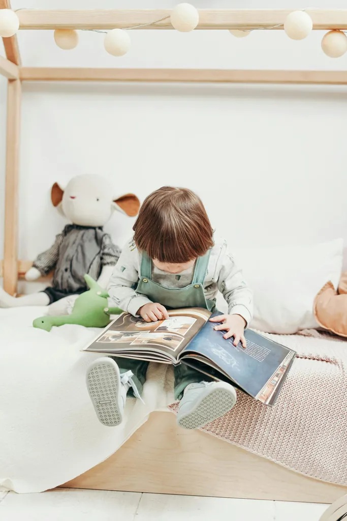 Un petit garçon assis sur un lit cabane feuillette un grand livre illustré, une peluche à côté de lui