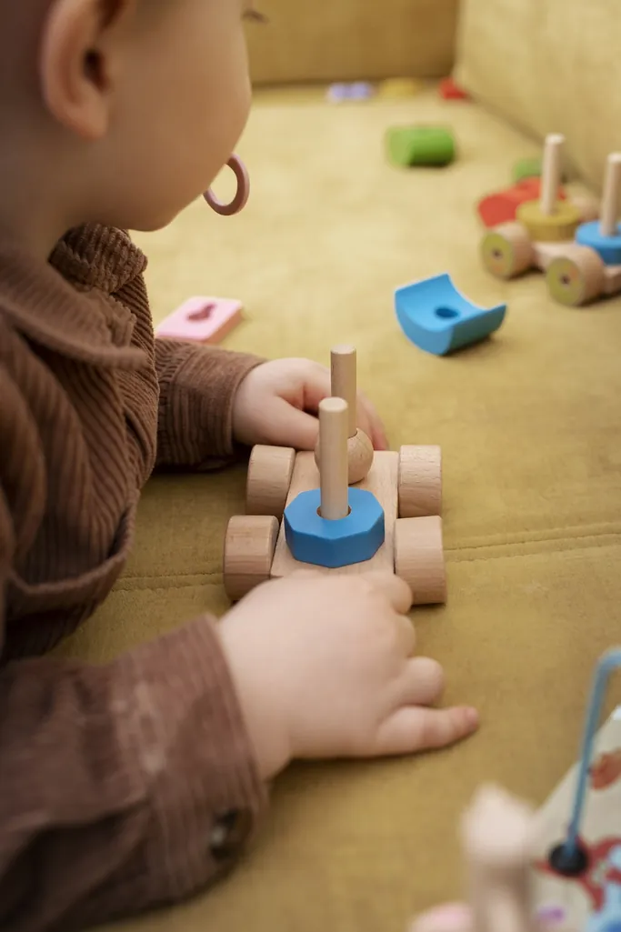 Un enfant en pull marron assemble les pièces d'un petit chariot en bois à empiler, sur un canapé jaune moutarde