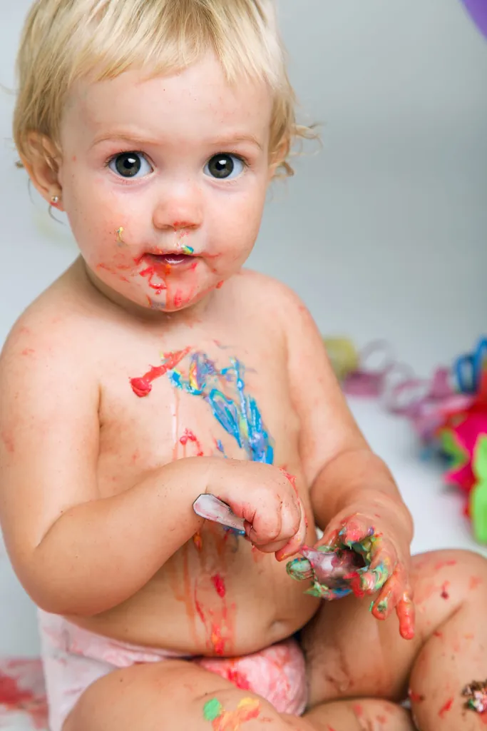 Une petite fille blonde, le visage et le ventre barbouillés de peinture de toutes les couleurs, les mains dans la peinture