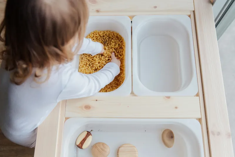 Un jeune enfant de dos qui plonge la main dans un bac rempli de pâtes, sur une table en bois à plusieurs bacs