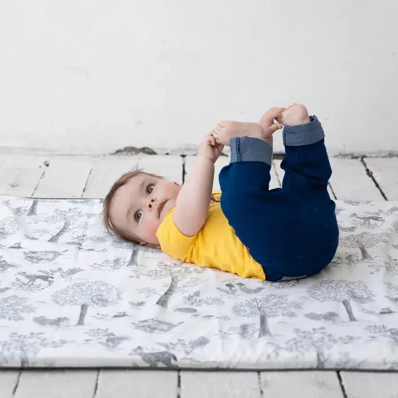 Un bébé en t-shirt jaune allongé sur le dos sur un tapis d’éveil gris clair imprimé d’arbres, qui attrape ses pieds
