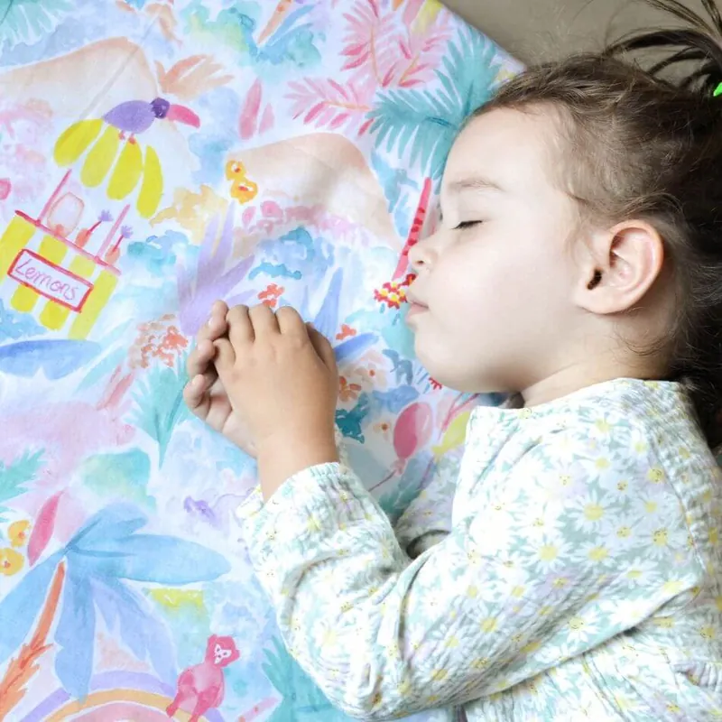 Une petite fille endormie sur un tapis imprimé de jungle aux couleurs pastel, les mains jointes sous la joue