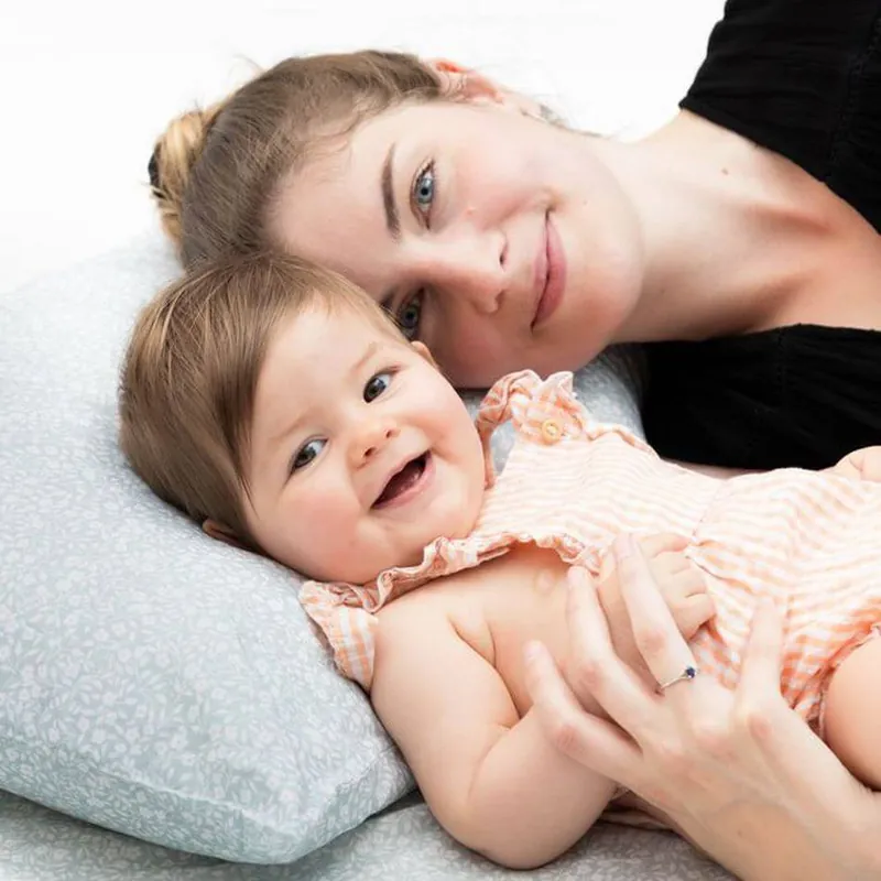 Une maman allongée joue contre joue avec sa petite fille souriante, sur un coussin au tissu gris clair