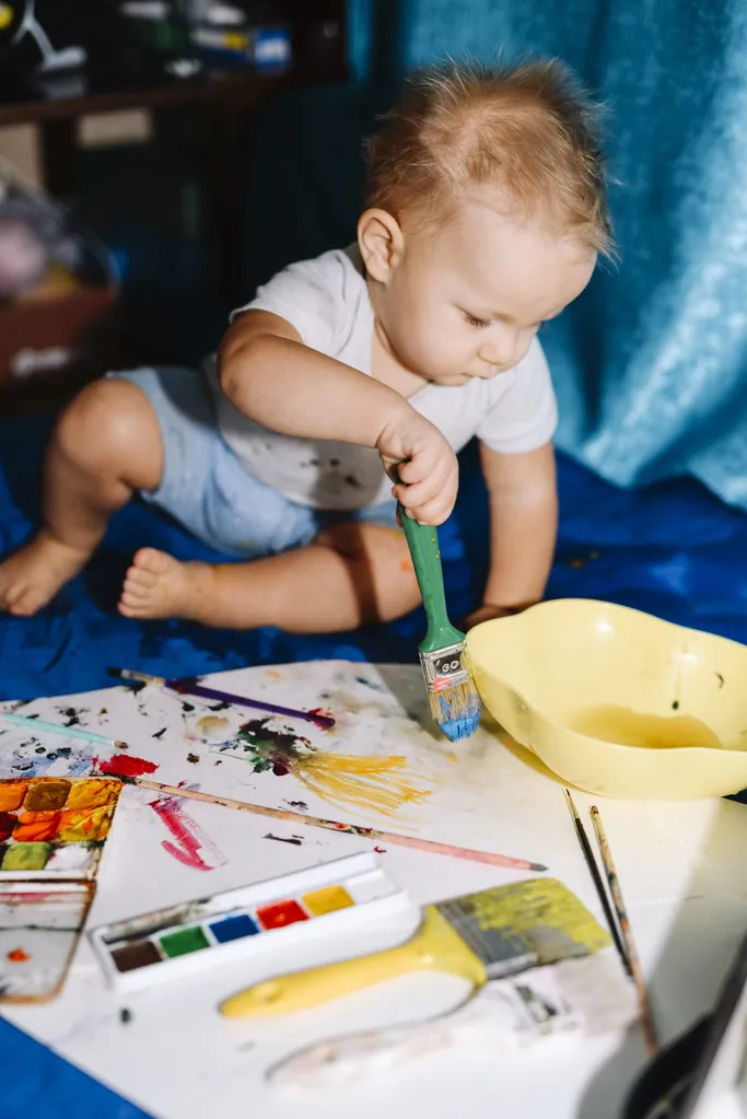 Un bébé assis qui trempe un pinceau dans la peinture, entouré de pinceaux, d’une boîte d’aquarelle et de feuilles peintes