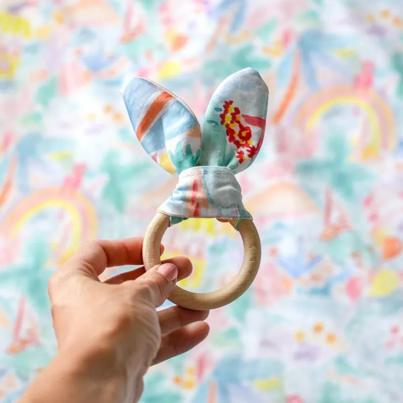 Une main qui tend un anneau de dentition en bois noué d’oreilles de lapin en tissu imprimé, devant un tapis aux couleurs pastel
