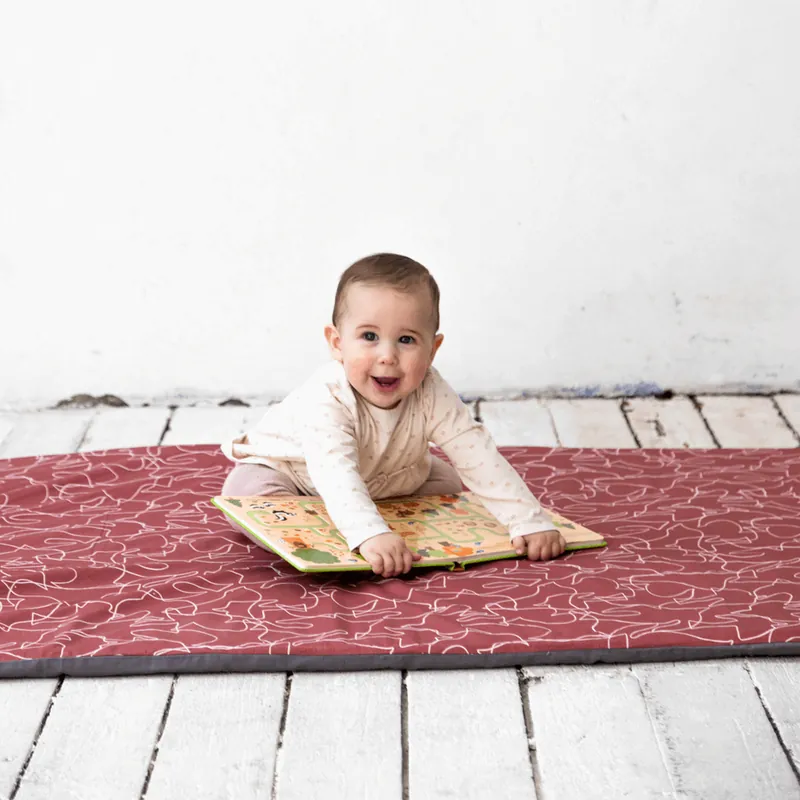 Un bébé souriant à plat ventre sur un tapis rouge brique posé sur un parquet blanc, les mains sur un livre illustré