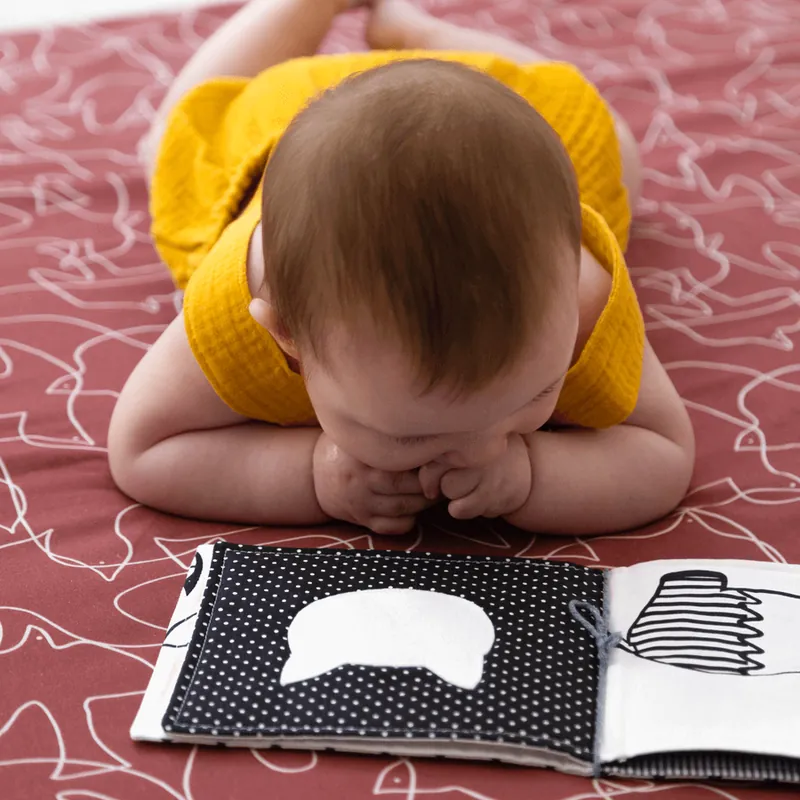 Un bébé en haut jaune, à plat ventre sur un tapis rouge brique, qui regarde un livre en tissu noir et blanc