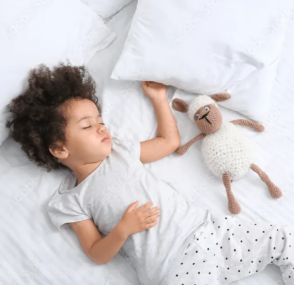 Une petite fille endormie sur le dos dans un lit blanc, à côté d'un mouton en crochet