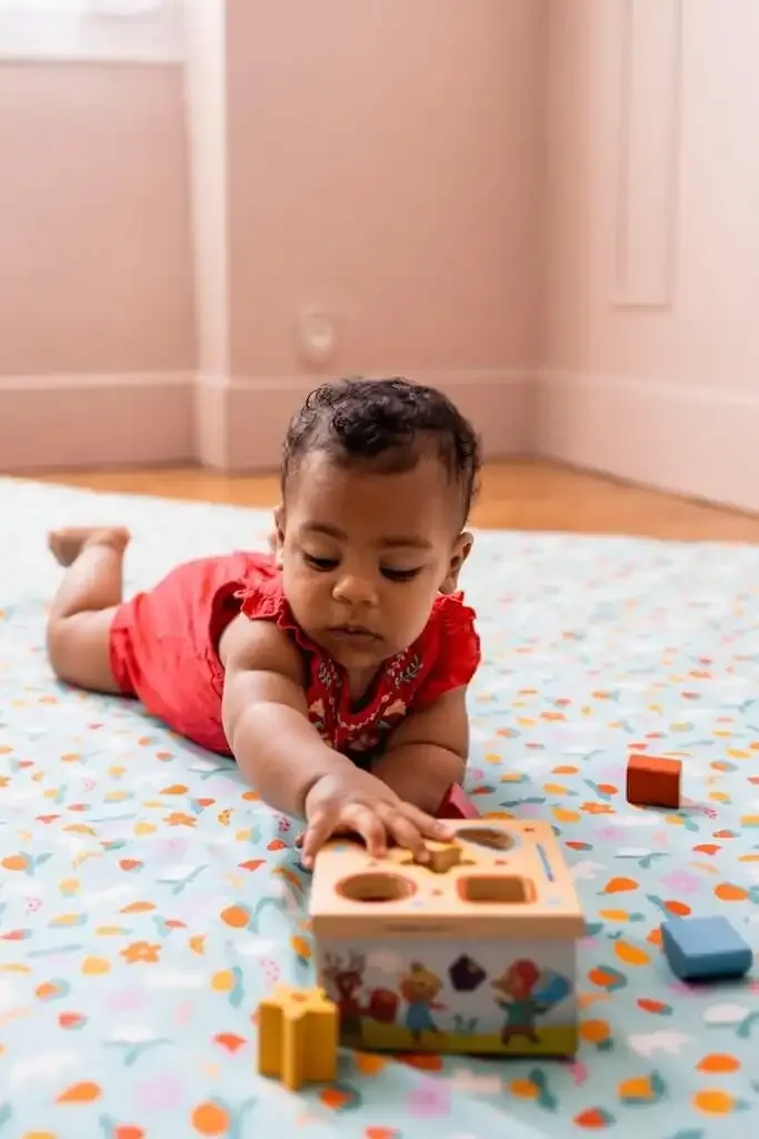 Un bébé en rouge, à plat ventre sur un tapis vert d'eau, tend la main vers une boîte à formes en bois