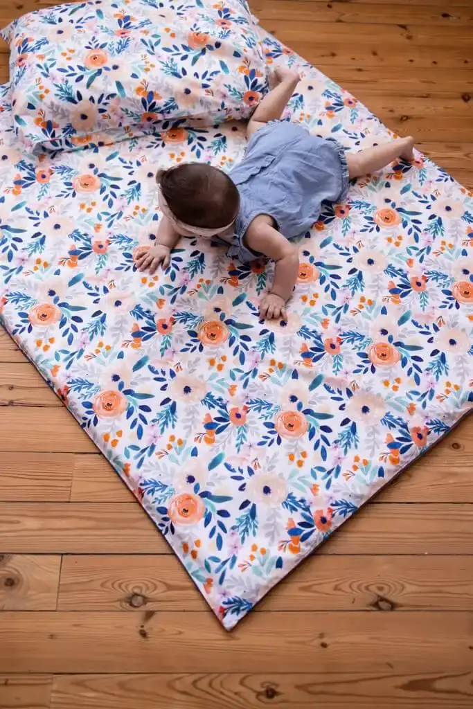 Un bébé en barboteuse en jean, à plat ventre sur un tapis d'éveil à fleurs posé sur un parquet, un coussin assorti derrière lui