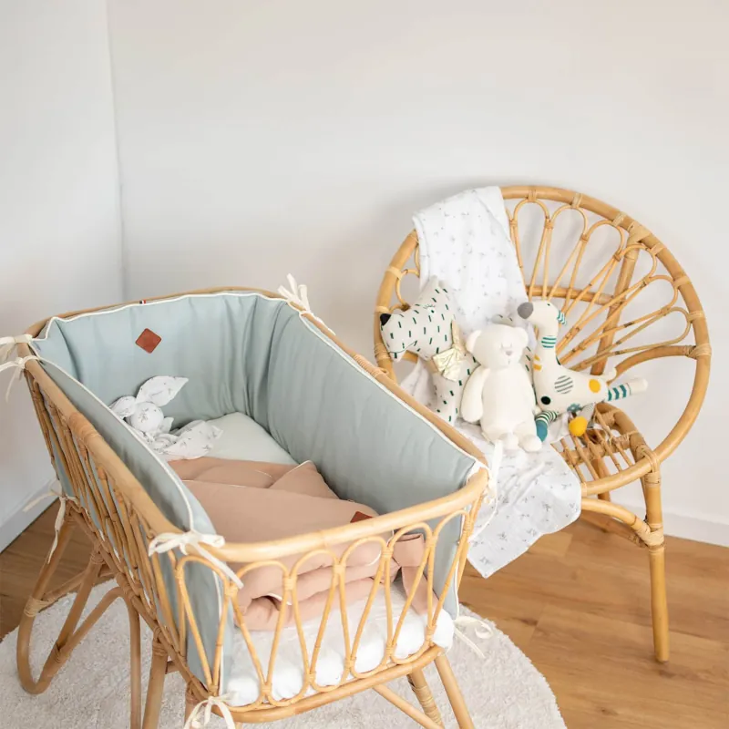 Un berceau en rotin garni de linge vert d'eau et de peluches, à côté d'un fauteuil en rotin, dans une chambre claire