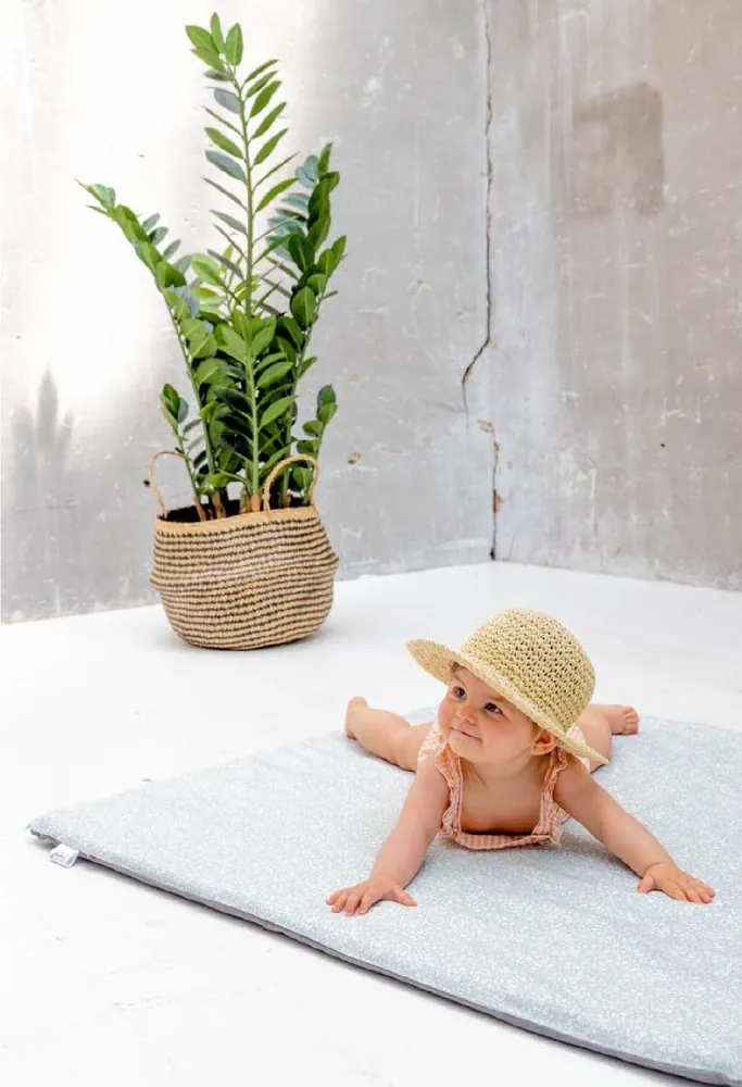 Un bébé coiffé d'un chapeau de paille, à plat ventre sur un tapis d'éveil gris clair, à côté d'une plante en pot