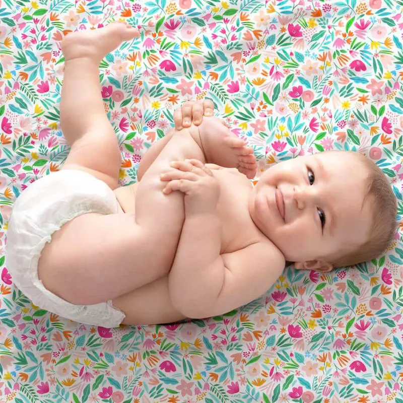 Un bébé souriant en couche, allongé sur le dos sur un tissu imprimé de fleurs colorées, qui attrape son pied