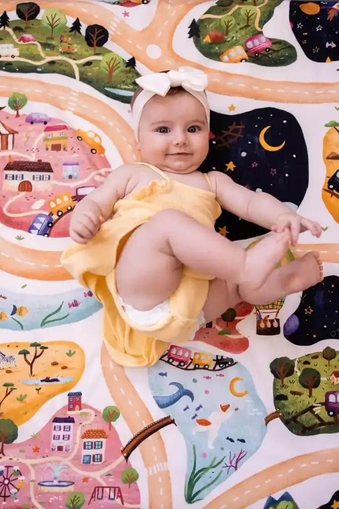 Un bébé coiffé d'un bandeau blanc, en body jaune, allongé sur le dos sur un tapis d'éveil imprimé de routes, de maisons et de forêts