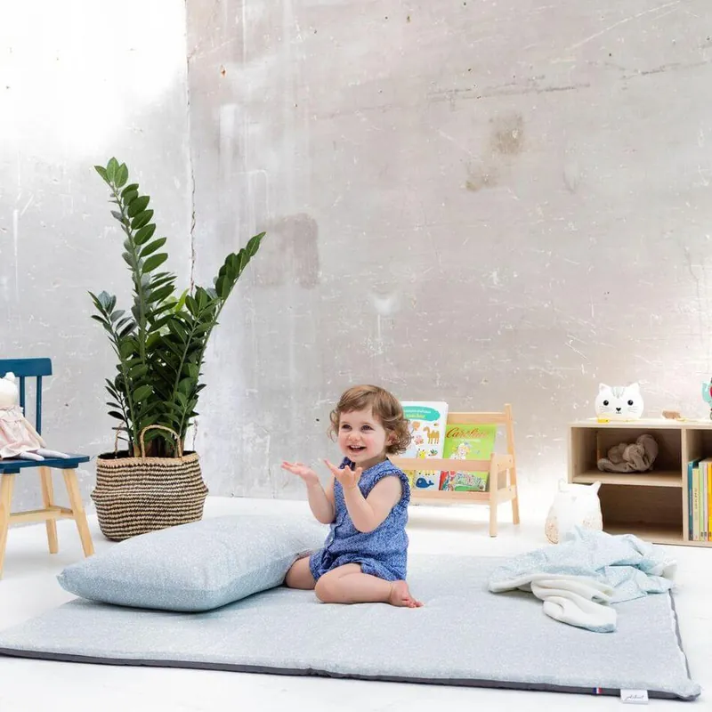 Une petite fille en barboteuse bleue, assise sur un tapis d'éveil gris clair, qui tape dans ses mains : aller à l'accueil de Jolihuit
