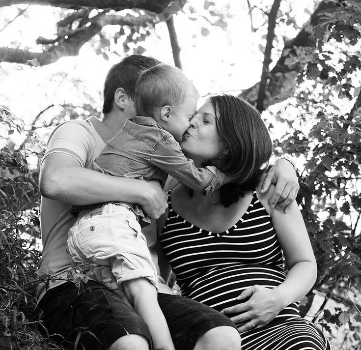 Photo en noir et blanc : un petit garçon dans les bras de son père embrasse sa mère enceinte, sous les arbres
