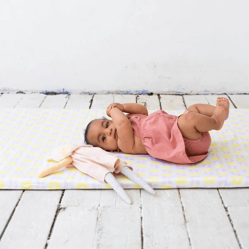Un bébé en barboteuse rose allongé sur le dos sur un tapis d'éveil à carreaux jaunes et lilas, à côté d'une peluche lapin