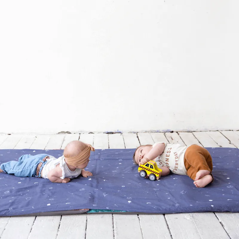 Deux bébés à plat ventre sur un tapis d'éveil bleu nuit étoilé posé sur un parquet blanc, l'un tenant une petite voiture jaune