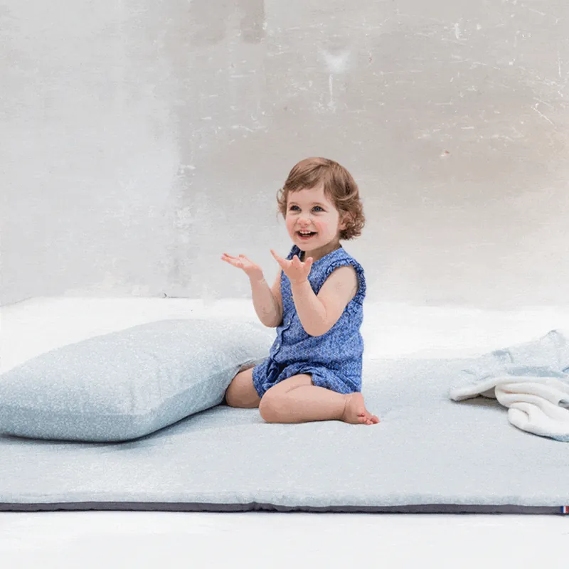Une petite fille en barboteuse bleue, assise sur un tapis d'éveil gris clair, qui tape dans ses mains : découvrir le tapis magique
