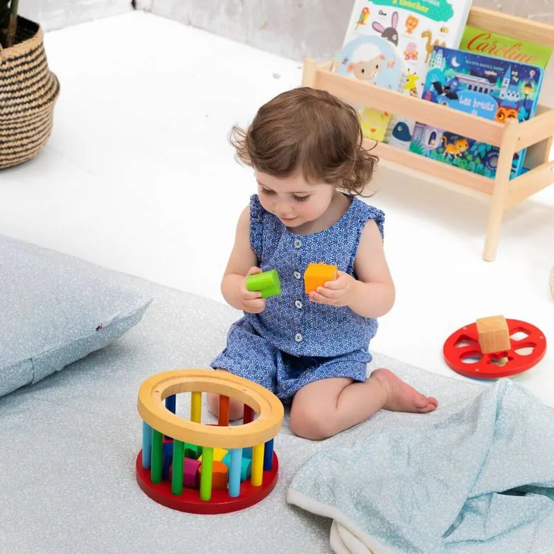 Une petite fille en barboteuse bleue, assise sur un tapis d'éveil gris clair, qui joue avec des jouets en bois devant une bibliothèque basse