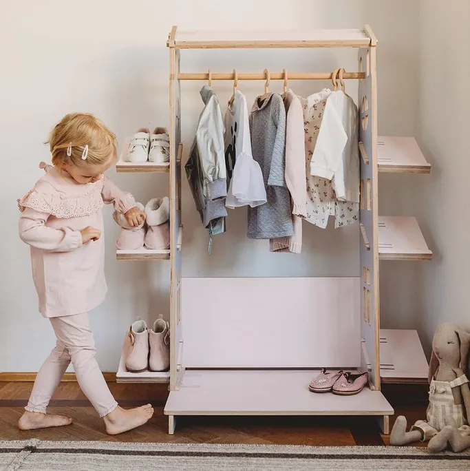 Une petite fille qui choisit ses chaussures dans une penderie basse en bois, avec vêtements suspendus et étagères à chaussures
