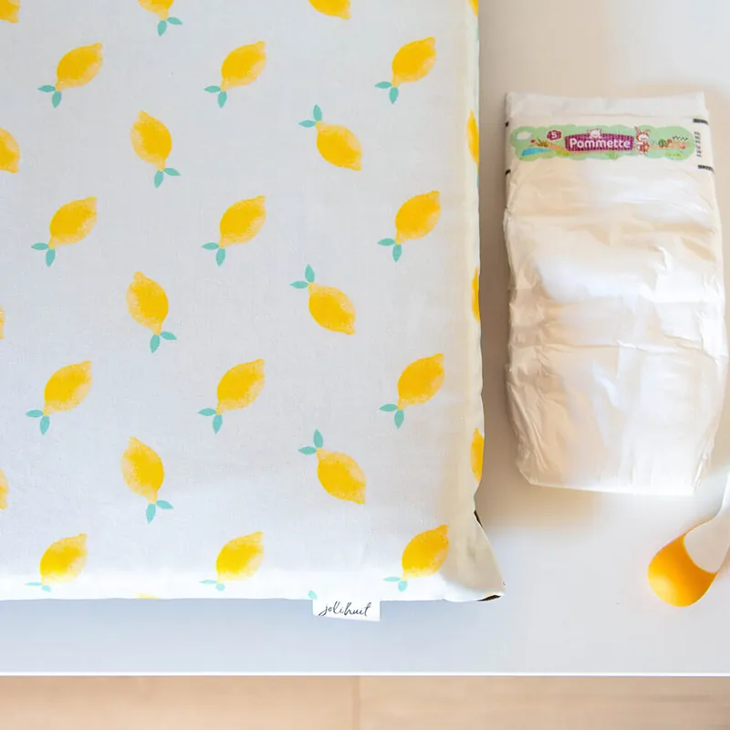 Un matelas à langer imprimé de citrons jaunes posé sur un meuble blanc, à côté d'un paquet de couches