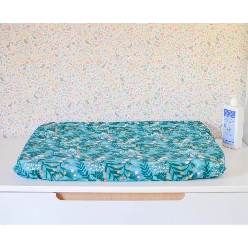 Un matelas à langer imprimé de feuillages sur fond bleu canard, posé sur un meuble blanc devant un papier peint fleuri, à côté d'un flacon de liniment