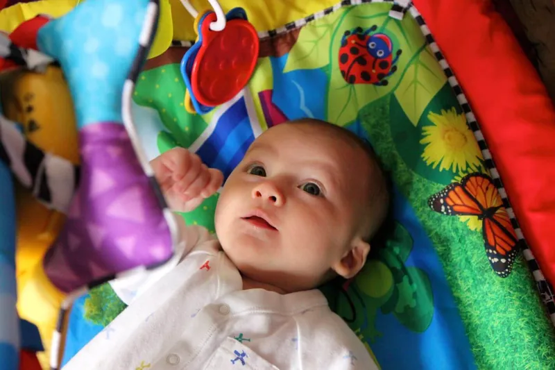 Un bébé allongé sur le dos sous une arche de jeu, sur un tapis d'éveil très coloré avec des jouets suspendus