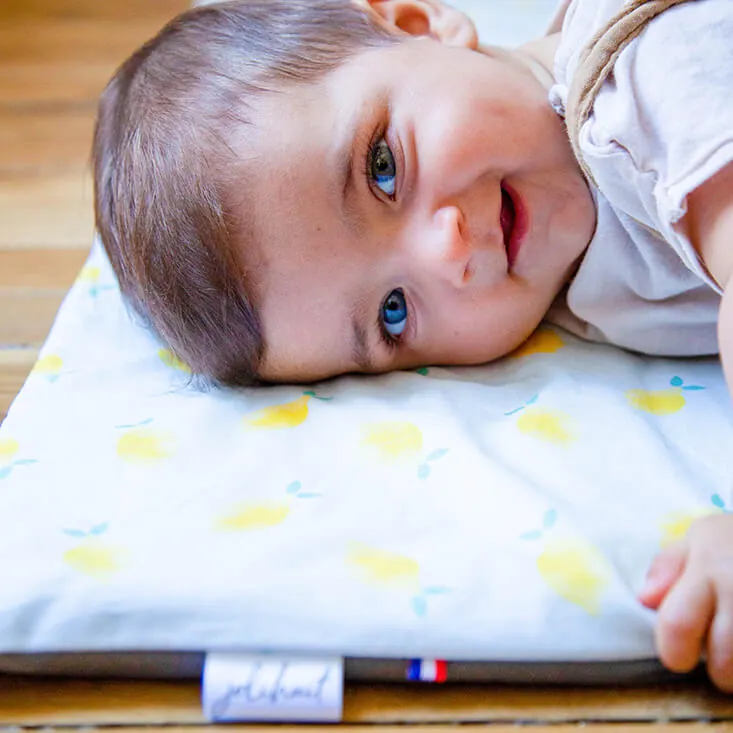 Un bébé souriant, la joue posée sur un tapis d'éveil blanc imprimé de citrons, l'étiquette Jolihuit au bord