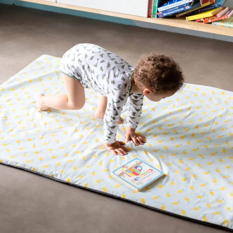 Un enfant à quatre pattes sur un grand tapis d'éveil blanc imprimé de citrons, près d'un livre, devant une bibliothèque