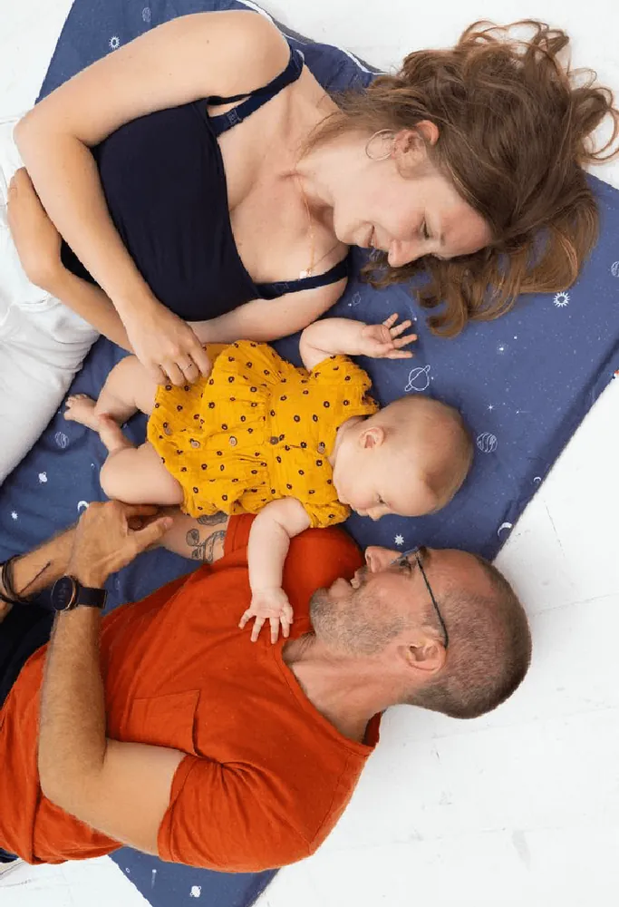 Un père et une mère allongés au sol de part et d'autre de leur bébé, sur un tapis d'éveil bleu nuit