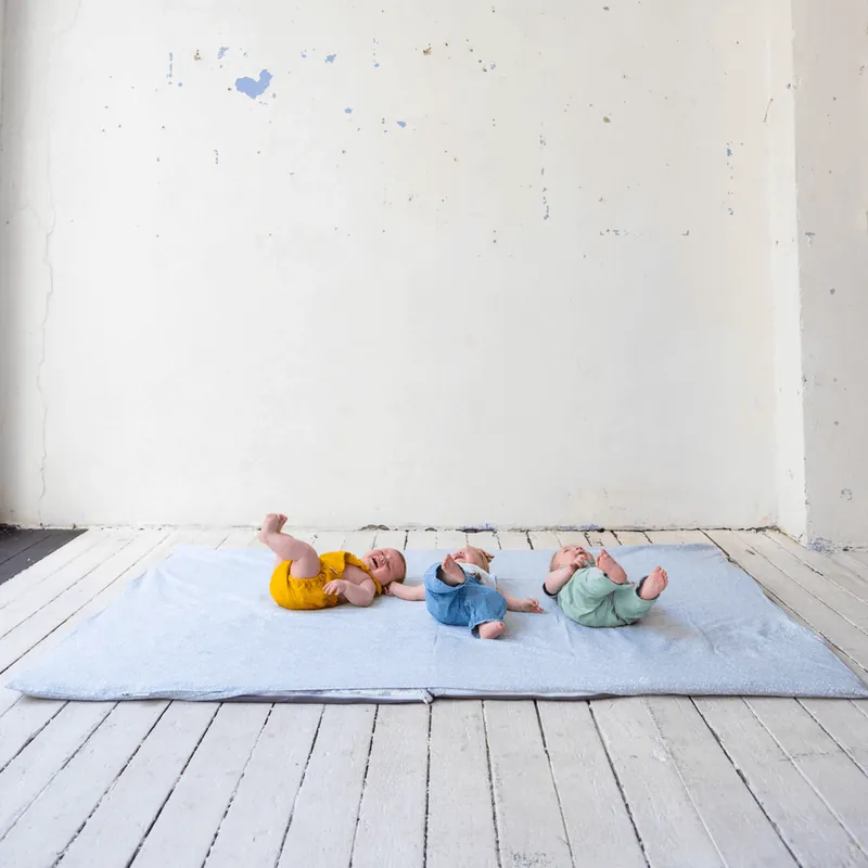 Trois bébés allongés côte à côte sur un grand tapis d'éveil bleu clair, posé sur un plancher blanc