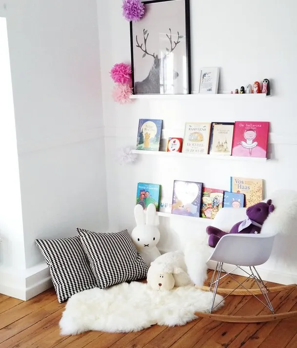Un coin lecture dans l'angle d'une chambre blanche : des livres posés face visible sur des étagères murales, des coussins, un fauteuil à bascule et des peluches sur une peau de mouton