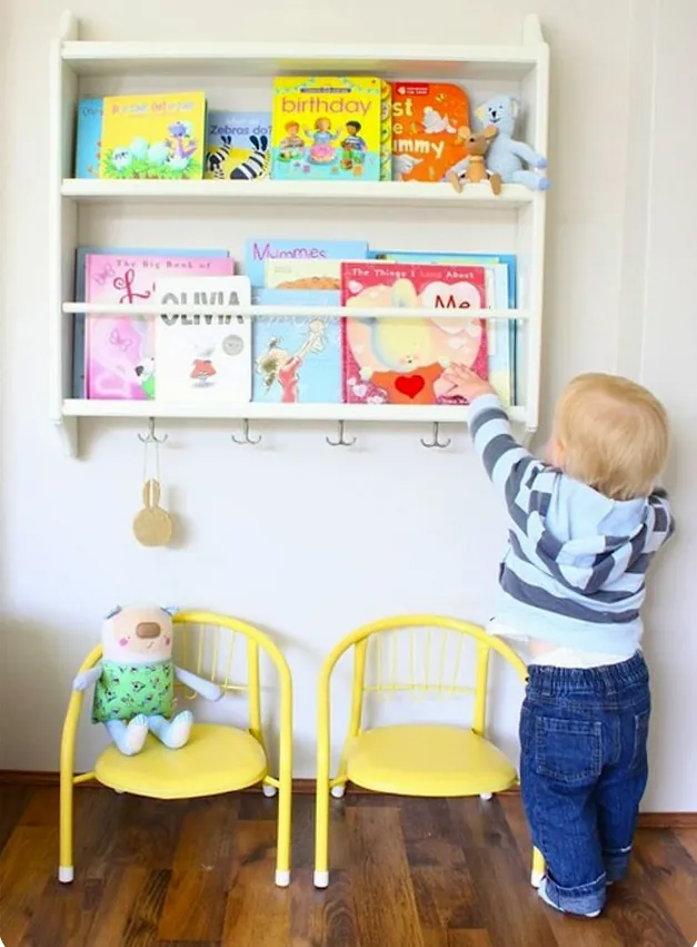 Un petit garçon debout qui attrape un livre sur une étagère murale blanche aux livres rangés face visible, au-dessus de deux petites chaises jaunes