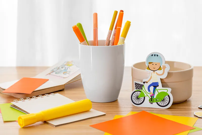 Un bureau d'enfant en bois avec un pot de feutres, un carnet, des papiers de couleur et une figurine en carton d'enfant à vélo