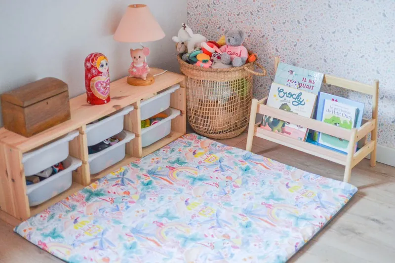Un coin lecture au sol : un tapis d'éveil imprimé pastel devant une bibliothèque basse en bois garnie de livres, des bacs de rangement et un panier de peluches