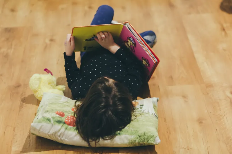 Un enfant allongé sur le ventre sur un parquet, la tête sur un coussin, qui lit un grand livre illustré