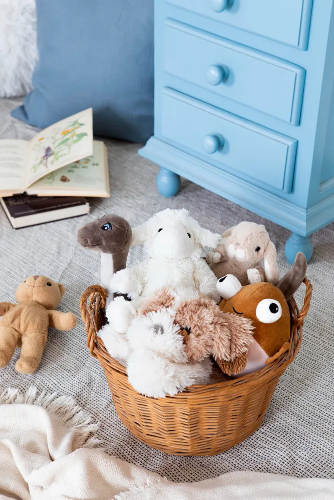 Un panier en osier rempli de peluches posé sur un tapis, devant une commode bleu ciel, à côté de livres ouverts